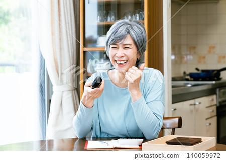 A senior woman happily watches television with the remote control in her hand. 140059972