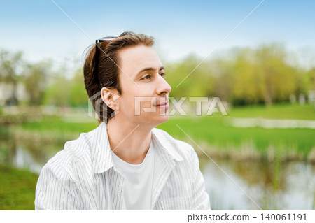 Close-up portrait of a young man enjoying a peaceful, sunny day in a park near a lake, looking away with a thoughtful and relaxed expression. Close-up portrait of a young man enjoying a peaceful, sunny day in a park near a lake, looking away with a thoughtful and relaxed expression. 140061191