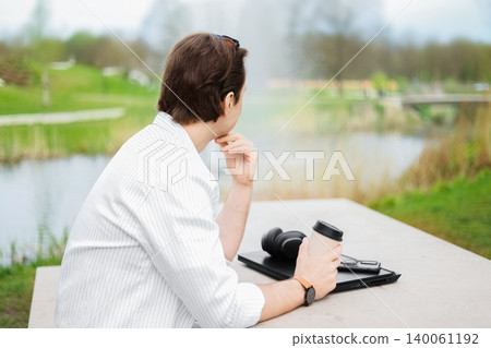 Back view of a freelance man relaxing in an outdoor park. He is taking a break from laptop work, holding a coffee and thinking while looking at nature. Back view of a freelance man relaxing in an outdoor park. He is taking a break from laptop work, holding a coffee and thinking while looking at nature. 140061192