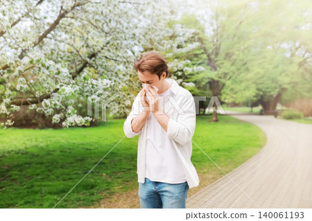 Man blowing his nose into a tissue in a park with spring blooms. Concept of hayfever, pollen allergy season, and healthcare. Man blowing his nose into a tissue in a park with spring blooms. Concept of hayfever, pollen allergy season, and healthcare. 140061193