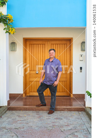 A man who owns a large house stands near the front door against the backdrop of a green garden 140061538