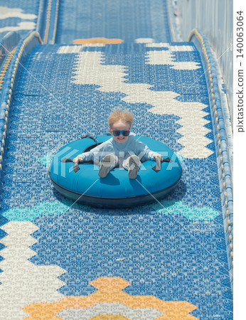 Blonde child in sunglasses rides a blue inflatable tube down an outdoor dry slide on a bright day Blonde child in sunglasses rides a blue inflatable tube down an outdoor dry slide on a bright day 140063064