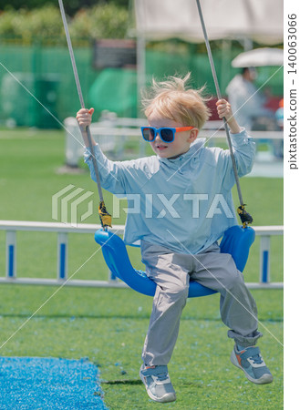 Smiling blond boy wearing blue sunglasses enjoying a sunny day swinging at the outdoor playground Smiling blond boy wearing blue sunglasses enjoying a sunny day swinging at the outdoor playground 140063066