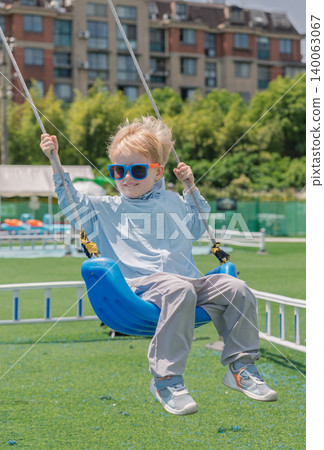 Smiling blond boy wearing blue sunglasses enjoying a sunny day swinging at the outdoor playground Smiling blond boy wearing blue sunglasses enjoying a sunny day swinging at the outdoor playground 140063067