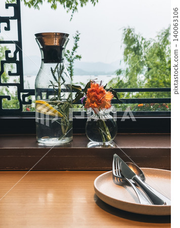 Dining table by window features refreshing water, rosemary, lemon, and flower with a beautiful lake view 140063106