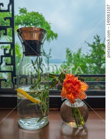 Clear carafe with lemon rosemary water and orange flowers decorating a wooden windowsill overlooking a serene lake 140063107