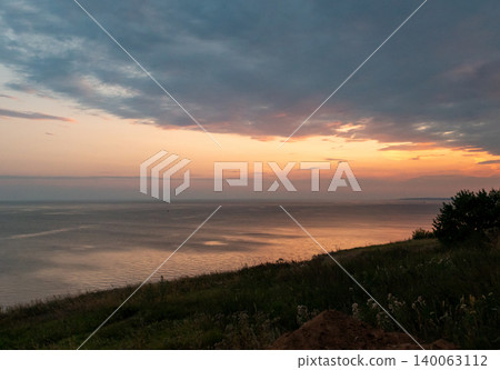 Vast water surface reflects vibrant sunset sky from a grassy hillside with trees during evening 140063112