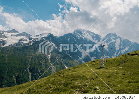 Cable car pylon stands on a green alpine slope with a dramatic backdrop of snow-capped mountains Cable car pylon stands on a green alpine slope with a dramatic backdrop of snow-capped mountains 140063934