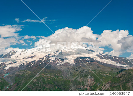 Mount Elbrus majestic peak reveals snow and glaciers contrasting with green lower slopes under a vibrant blue sky Mount Elbrus majestic peak reveals snow and glaciers contrasting with green lower slopes under a vibrant blue sky 140063947