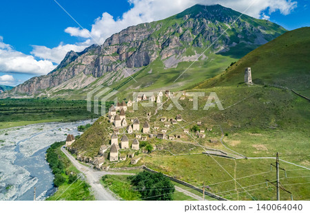 Aerial view captures Dargavs City of the Dead sit on a hill near a river with mountains in the background during a sunny day in North Ossetia-Alania 140064040