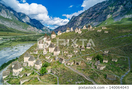Aerial view captures Dargavs City of the Dead sit on a hill near a river with mountains in the background during a sunny day in North Ossetia-Alania 140064043