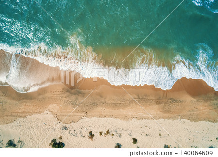 Aerial view shows vibrant green ocean water with white foam waves crashing onto a tan sandy beach. Aerial view shows vibrant green ocean water with white foam waves crashing onto a tan sandy beach. 140064609