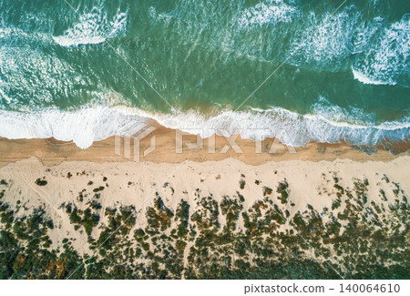 Aerial view shows vibrant green ocean water with white foam waves crashing onto a tan sandy beach. Aerial view shows vibrant green ocean water with white foam waves crashing onto a tan sandy beach. 140064610