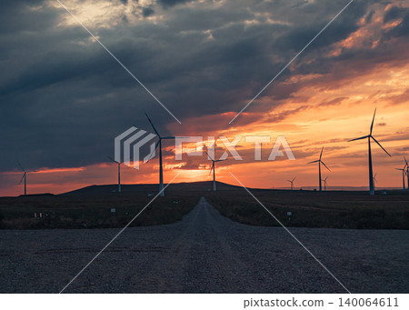 Wind turbines stand against a vibrant sunset sky above a rural landscape 140064611