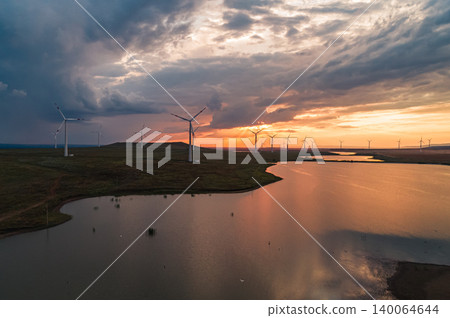 Aerial view of a wind turbines generate clean energy during sunset over a tranquil lake in a rural landscape with open fields Aerial view of a wind turbines generate clean energy during sunset over a tranquil lake in a rural landscape with open fields 140064644
