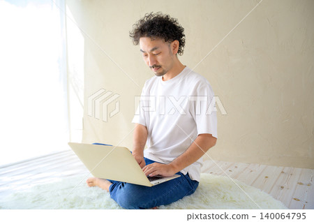A smiling Japanese man using a laptop at home. A smiling Japanese man using a laptop at home. 140064795