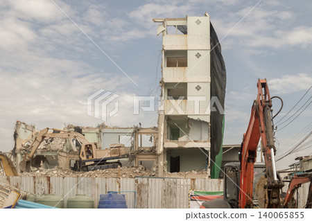 a slender, multi-story building stands partially exposed under a cloud-dappled sky, draped in a dark safety net while heavy excavators systematically reduce the surrounding architecture to a landscape a slender, multi-story building stands partially exposed under a cloud-dappled sky, draped in a dark safety net while heavy excavators systematically reduce the surrounding architecture to a landscape 140065855