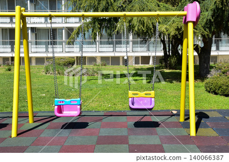 Brightly colored swings with yellow frames and pink seats are positioned in a grassy area of an urban park, surrounded by trees and a building in the background, creating a playful atmosphere Brightly colored swings with yellow frames and pink seats are positioned in a grassy area of an urban park, surrounded by trees and a building in the background, creating a playful atmosphere 140066387