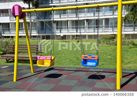 Brightly colored swings with yellow frames and pink seats are positioned in a grassy area of an urban park, surrounded by trees and a building in the background, creating a playful atmosphere Brightly colored swings with yellow frames and pink seats are positioned in a grassy area of an urban park, surrounded by trees and a building in the background, creating a playful atmosphere 140066388