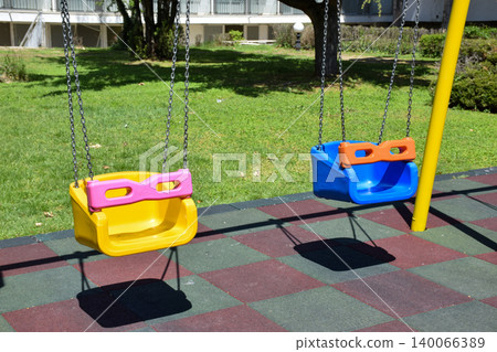 Brightly colored swings with yellow frames and pink seats are positioned in a grassy area of an urban park, surrounded by trees and a building in the background, creating a playful atmosphere Brightly colored swings with yellow frames and pink seats are positioned in a grassy area of an urban park, surrounded by trees and a building in the background, creating a playful atmosphere 140066389