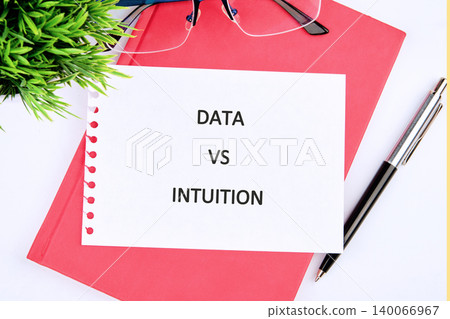 A white paper displays DATA VS INTUITION atop a red notebook, accompanied by a pen, eyeglasses, and a potted plant, illustrating the critical balance in modern decision making 140066967