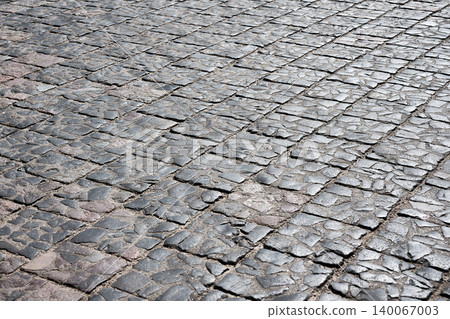Bright sunlight illuminates an ancient cobblestone street. Rough, irregular grey stones form a weathered, textured ground pattern. The sturdy paving shows signs of age 140067003