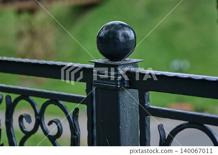 A dark wrought iron fence post featuring an ornamental sphere top and intricate scroll design stands outdoors A dark wrought iron fence post featuring an ornamental sphere top and intricate scroll design stands outdoors 140067011