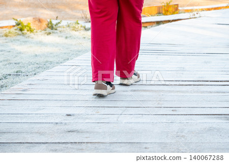 Woman walking on wooden boardwalk covered with frost. Girl in red trousers exploring nature during a cold winter morning. Outdoor activity, healthy lifestyle and seasonal travel concept. Woman walking on wooden boardwalk covered with frost. Girl in red trousers exploring nature during a cold winter morning. Outdoor activity, healthy lifestyle and seasonal travel concept. 140067288