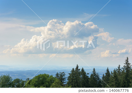 Large fluffy cumulus cloud in a blue sky over a distant hazy landscape. Dark green coniferous trees in the foreground. Natural weather background with copy space 140067776
