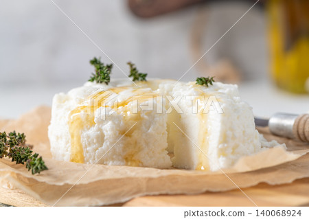 A close up shot of a wheel of fresh white ricotta cheese drizzled with golden honey and garnished with sprigs of aromatic thyme resting on brown parchment paper over a wooden board in a bright kitchen A close up shot of a wheel of fresh white ricotta cheese drizzled with golden honey and garnished with sprigs of aromatic thyme resting on brown parchment paper over a wooden board in a bright kitchen 140068924
