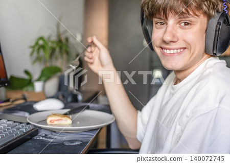 Smiling teenage gamer wearing headset and eating sweet pastry from a plate while sitting at the PC desk at home. Smiling teenage gamer wearing headset and eating sweet pastry from a plate while sitting at the PC desk at home. 140072745