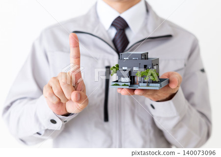 一位身穿灰色工作服的中年商人舉起一個房屋模型,在白色背景下豎起食指。 一位身穿灰色工作服的中年商人舉起一個房屋模型,在白色背景下豎起食指。 140073096