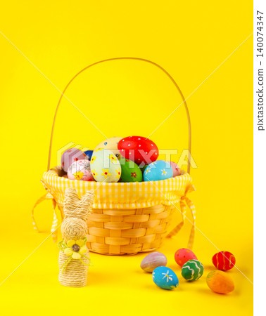 A festive vertical shot of a wicker basket overflowing with colorful hand-painted Easter eggs and a straw bunny figurine, set against a bright, solid yellow background. High quality photo A festive vertical shot of a wicker basket overflowing with colorful hand-painted Easter eggs and a straw bunny figurine, set against a bright, solid yellow background. High quality photo 140074347