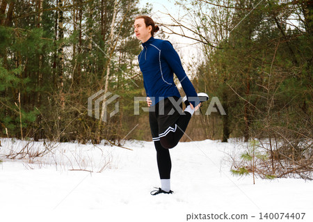 Male runner doing a quad stretch in a snowy forest. Active young man warming up for a winter outdoor workout in athletic sportswear. High quality photo 140074407
