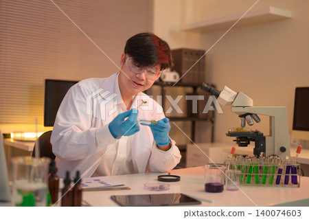 Scientist carefully examines a plant sample with tweezers beside a microscope Scientist carefully examines a plant sample with tweezers beside a microscope 140074603