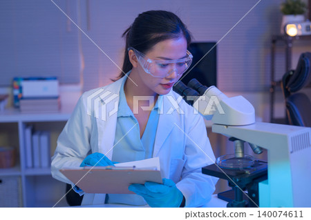 Female scientist using a microscope to analyze samples and writing on document Female scientist using a microscope to analyze samples and writing on document 140074611