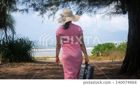 Young woman in pink dress and straw hat carrying a suitcase walking toward the sea visible ahead through tropical trees. Beach vacation concept 140074668