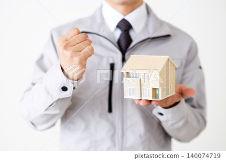A middle-aged businessman in gray work clothes strikes a triumphant pose while holding a model of a house, against a white background. A middle-aged businessman in gray work clothes strikes a triumphant pose while holding a model of a house, against a white background. 140074719