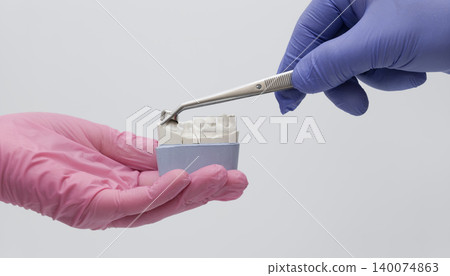 Hands in blue and pink gloves fitting a metal dental crown onto a plaster jaw model using tweezers. Isolated on white. Hands in blue and pink gloves fitting a metal dental crown onto a plaster jaw model using tweezers. Isolated on white. 140074863