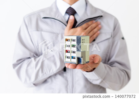 A middle-aged businessman in gray work clothes holding a model of an apartment building against a white background. A middle-aged businessman in gray work clothes holding a model of an apartment building against a white background. 140077072