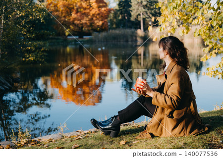 Woman sits holding cup on grassy bank by pond in autumn park. Mindful recreation by water, outdoor wellness lakeside destination, mental wellbeing nature connection. Woman sits holding cup on grassy bank by pond in autumn park. Mindful recreation by water, outdoor wellness lakeside destination, mental wellbeing nature connection. 140077536