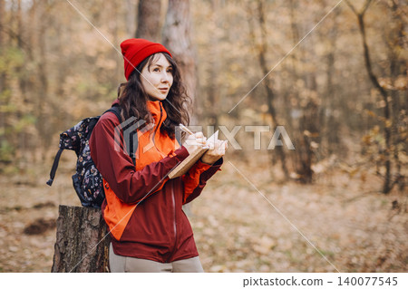 Woman stands in autumn forest while writing in notebook and looking away. Wilderness survival curriculum, nature-based learning program, forest school activities. Woman stands in autumn forest while writing in notebook and looking away. Wilderness survival curriculum, nature-based learning program, forest school activities. 140077545