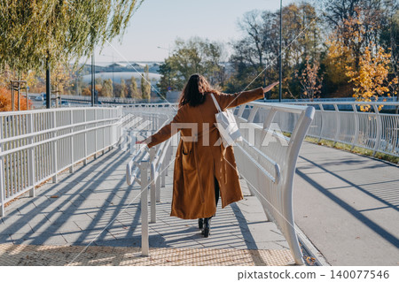 Woman walks with outstretched arms on pedestrian bridge with back to camera. Solo female urban travel, exploring modern city destinations, autumn city break tour. Woman walks with outstretched arms on pedestrian bridge with back to camera. Solo female urban travel, exploring modern city destinations, autumn city break tour. 140077546
