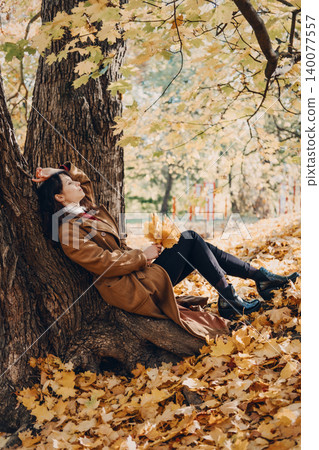 Woman rests against large tree trunk in autumn forest holding leaves with closed eyes. Grounding in nature autumn, Sensory Walking, Physical Earthing, Cycle of Letting Go. Woman rests against large tree trunk in autumn forest holding leaves with closed eyes. Grounding in nature autumn, Sensory Walking, Physical Earthing, Cycle of Letting Go. 140077557