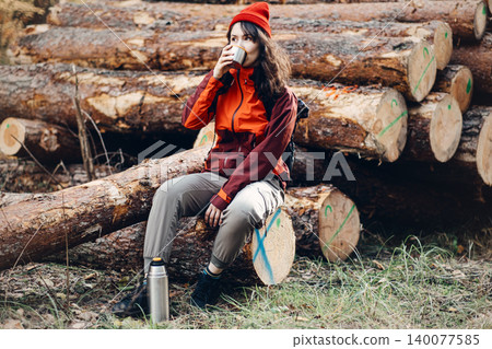 Woman drinks hot beverage while sitting on fallen logs in forest with metal thermos nearby. Cold weather hiking essentials, necessary outdoor equipment, thermos use in wilderness. Woman drinks hot beverage while sitting on fallen logs in forest with metal thermos nearby. Cold weather hiking essentials, necessary outdoor equipment, thermos use in wilderness. 140077585