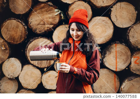 Smiling woman pours drink from metal thermos into cup while standing in front of stacked logs in forest. Cold weather hiking essentials, necessary outdoor equipment, thermos use in wilderness. Smiling woman pours drink from metal thermos into cup while standing in front of stacked logs in forest. Cold weather hiking essentials, necessary outdoor equipment, thermos use in wilderness. 140077588