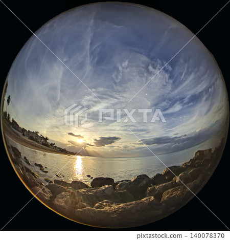 Sunset occurs over the sea with sun rays breaking through clouds. Waves gently lap against the rocks along the shore. The sky is filled with colors and formations from the clouds. Sunset occurs over the sea with sun rays breaking through clouds. Waves gently lap against the rocks along the shore. The sky is filled with colors and formations from the clouds. 140078370