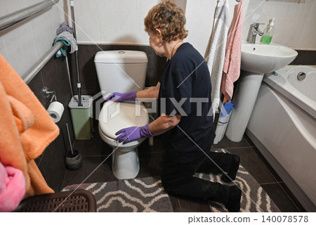 Woman cleaning toilet in a small bathroom during daytime hours Woman cleaning toilet in a small bathroom during daytime hours 140078578
