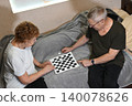 Elderly couple plays checkers together on a gray couch in their living room 140078626