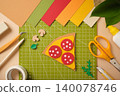 Flat lay of a paper pizza slice with pepperoni on a green cutting mat. Creative DIY workspace with colorful paper sheets, scissors, glue, tweezers, and stationery for art and craft projects. 140078746